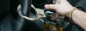 Man In Handcuffs Holds Bottle Of Alcohol In Car. Drunk Driving Concept. Don't Drink And Drive.